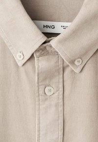 Camisa de pana beige con cuello puntiagudo, placket frontal con botones blancos y una etiqueta que dice "MNG EUR-USE ME."