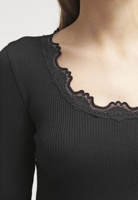 Musta ribattu toppi, jossa on kontrastivärinen musta pitsireunus. Tekstuuri on sileä ja muoto on istuva.