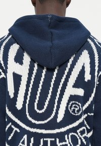 Marineblå strikket hettegenser med hvit grafisk design. Har et fremtredende logo, strukturert søm og en tettsittende hette. Ingen synlige lukninger.
