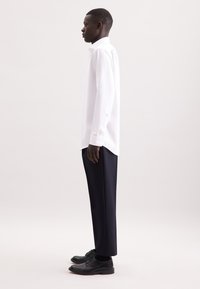 Camicia bianca a maniche lunghe, in cotone, con colletto button-down, abbinata a pantaloni eleganti blu navy e scarpe nere, vista di lato.