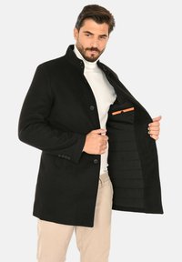 Cappotto nero in lana con colletto alla coreana, design monopetto e chiusura con bottoni. Interno foderato con tasca decorativa arancione.