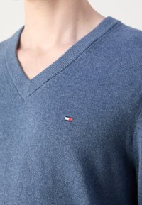 Gros plan d'une personne portant un pull bleu à col en V avec un petit écusson rectangulaire rouge, blanc et noir sur le côté gauche de la poitrine.