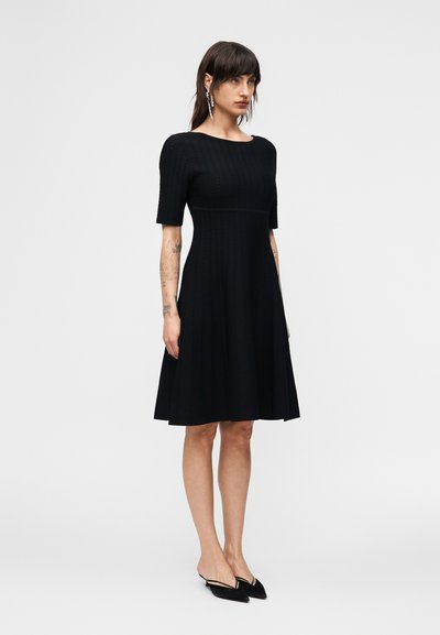 Γυναίκα που φοράει μαύρο φόρεμα μέχρι το γόνατο με κοντά μανίκια, μαύρες γόβες με λουράκι στο πίσω μέρος και μακριά σκουλαρίκια.