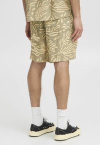 Shorts beiges avec un motif de feuilles, fabriqués en matériau léger, disposent de poches latérales et d'une coupe décontractée. Baskets noires avec des accents blancs.