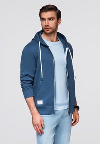 Niebieska bluza zapinana na suwak z białymi sznurkami, przednimi kieszeniami i ściągaczami, noszona na jasnoniebieskiej koszuli, w połączeniu z jasnymi dżinsami.