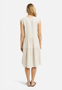 Frau steht mit dem Rücken zur Kamera, trägt ein �ärmelloses beiges Midikleid mit rüschigen Schultern und braunen flachen Sandalen vor einem weißen Hintergrund.