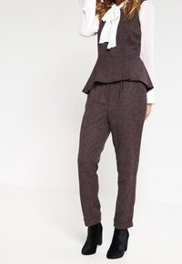 Monos de peplum marrón hechos de tela texturizada, con una blusa blanca de lazo debajo y pantalones ajustados y ceñidos. Botas negras hasta el tobillo visibles.