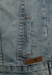 Ljusblå denimtyg med synlig sömnad; har en liten ficka och en brun läderlapp med en stjärndetalj.