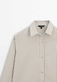 Randig skjorta i vitt och ljusbrunt, med en klassisk krage, knäppt fram och slät textur. Inkluderar två knappar och tunna ränder.