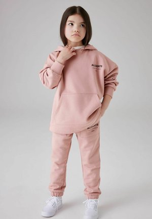 Felpa rosa chiaro con cappuccio e tasca frontale, abbinata a pantaloni jogging coordinati. Entrambi presentano accenti di logo neri. Sneakers bianche completano il look.