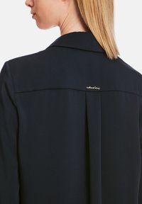 Marinebluse mit strukturiertem Kragen, Faltendetail am Rücken und einem goldenen Akzent-Logo in der Nähe des Ausschnitts. Glatte Textur des Stoffes.