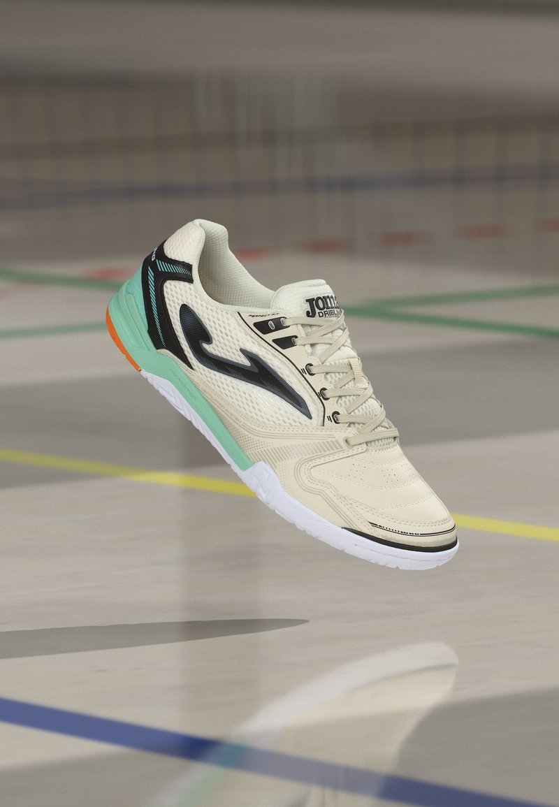 Valge sisejalats spordiks, millel on must logo, mündirohelised ja oranžid tallamustrid, paigutatud gymsaliku põranda kohale, millel on värvilised jooned.