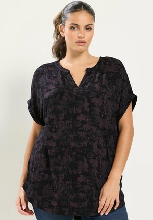 Femme aux cheveux foncés attachés en arrière, portant un haut noir ample à manches courtes avec des motifs floraux subtils et un jean bleu sur un fond uni.