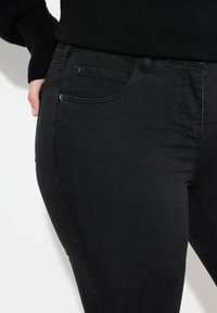 Jeans de mezclilla negra con una textura suave, ligero estiramiento, cinco bolsillos, bolsillos frontales en ángulo y detalles metálicos en la cintura.