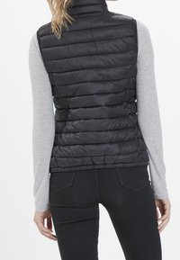 Černá prošívaná vesta s horizontálním prošitím, vysokým límcem a elastickým lemem. Nošena přes světle šedé tričko s dlouhým rukávem a upnuté černé kalhoty.