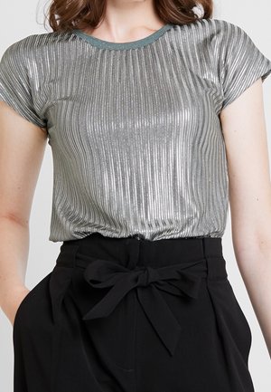 Vrouw draagt een zilverkleurige metallic geribde top met korte mouwen, ingestopt in een zwarte broek met hoge taille en een geknoopt stoffen riem.