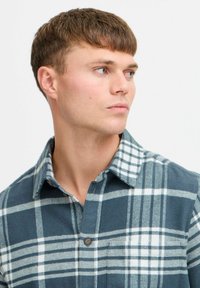 Checkered flannel shirt in dark teal with white patterns, button-up, long sleeves, collar, and soft texture. Features chest pocket detail.