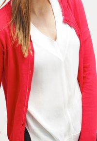 Cardigan rouge en maille légère, avec fermeture à boutons, poignets côtelés et design uni, porté sur un haut blanc.