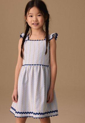 Niña joven con el pelo trenzado que lleva un vestido blanco sin mangas con rayas verticales azules y bordes con volantes, de pie frente a un fondo liso.