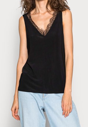 Kobieta w czarnej bluzce na ramiączkach z dekoltem w kształcie litery V wykończonym koronką oraz jasnoniebieskich dżinsach, stojąca na tle jednolitego tła.