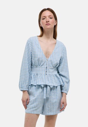 Femme portant un chemisier à manches longues à carreaux bleu et blanc avec des boutons et un short assorti, debout avec une expression neutre sur fond gris.