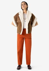 Veste en daim marron avec un col en shearling blanc, associée à un pantalon droit orange vif et des chaussures bourgogne. Accessoires simples inclus.