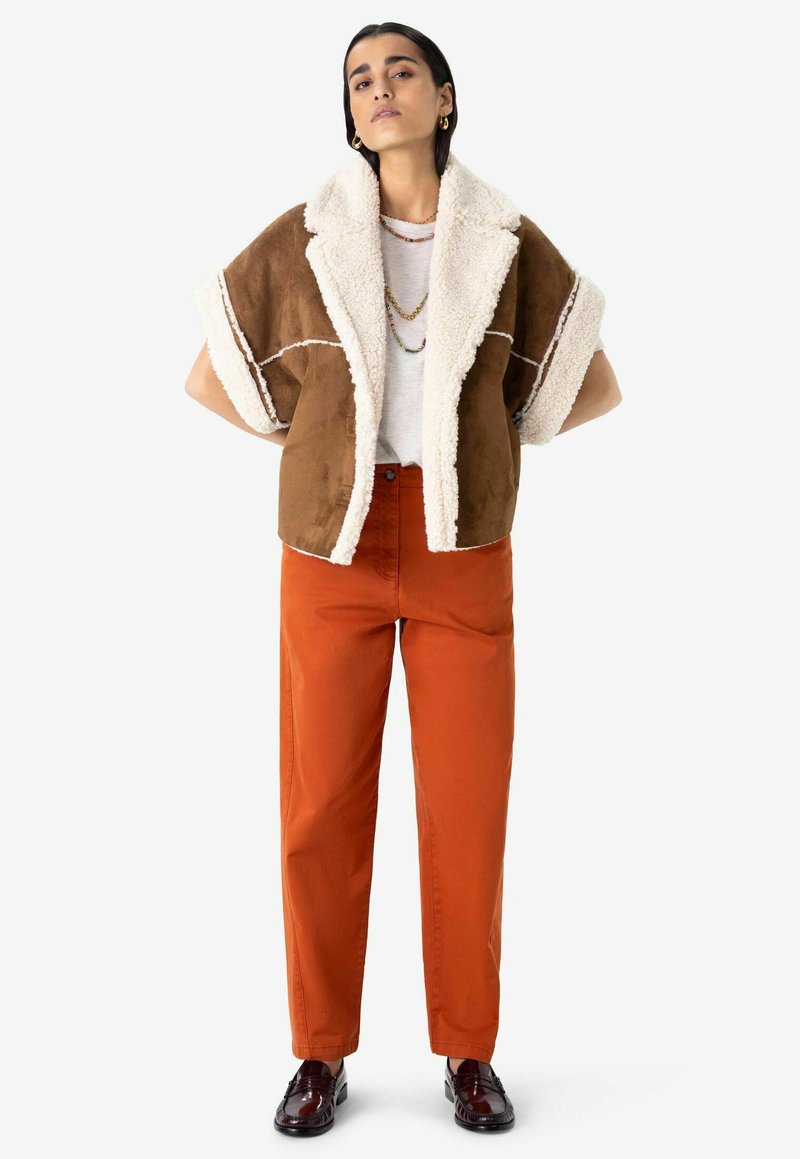 Veste en daim marron avec un col en shearling blanc, associée à un pantalon droit orange vif et des chaussures bourgogne. Accessoires simples inclus.