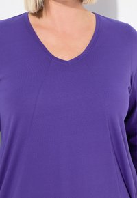 Lila V-Ausschnitt-Oberteil aus weichem Baumwollstoff. Mit langen Ärmeln und Nahtdetails auf der Vorderseite, die eine entspannte Passform bieten.