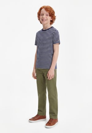 Smilende rødhåret dreng, der står, iført en marineblå og hvidstribet t-shirt, olivengrønne bukser og brune sneakers på hvid baggrund.