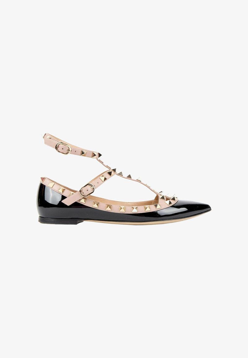 Chaussure plate en cuir verni noir avec des brides beige à la cheville ornées de clous pyramides dorés et deux boucles réglables.
