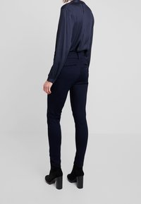 Kvinna som står vänd bort, klädd i slim-fit byxor i marinblått, marinblå långärmad blus och svarta klackade ankelskor mot en neutral bakgrund.