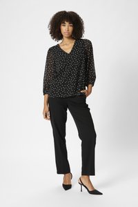 Blouse noire transparente à pois blancs, décolleté en V, et manches bouffantes 3/4, associée à un pantalon noir à jambes droites et des escarpins noirs à talons pointus.