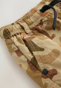 Pantaloni corti mimetici con cintura elastica, cordoncino e dettaglio a bottone. Il materiale presenta diverse tonalità di beige, marrone e modelli arancioni.