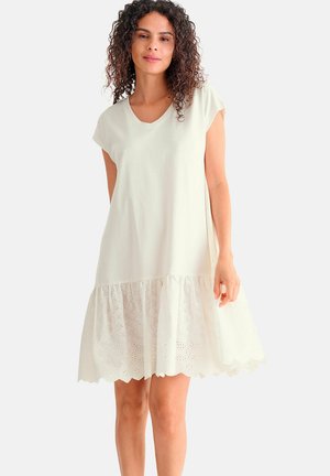 Femme aux cheveux bouclés portant une robe ample à manches courtes couleur crème avec un ourlet brodé en forme de festons, posant devant un fond uni.