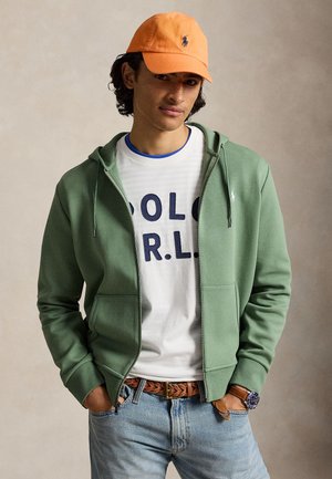 Jeune homme portant une casquette orange, un sweat à capuche zippé vert, un t-shirt blanc avec un motif graphique, une ceinture tressée, un jean bleu et une montre-bracelet, mains dans les poches.