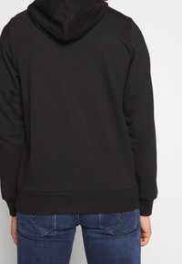 Sudadera con capucha negra de textura suave, capucha en la parte trasera y puños acanalados. Llevada sobre jeans azules con diseño clásico de cinco bolsillos.