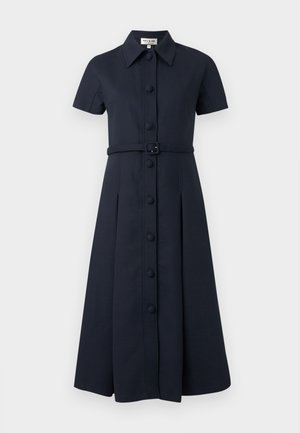 Γυναίκα που στέκεται με τα χέρια στις τσέπες, φοράει ένα μαύρο midi φόρεμα με κουμπιά και κοντά μανίκια και λευκά μυτερά παπούτσια.