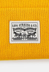 Gul strikket hue med ribbet tekstur og et hvidt mærke med sorte grafikker, der står "Levi Strauss & Co." og "Kvalitetsbeklædning."