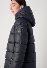 Vrouw met lang, donker haar draagt een donkere gewatteerde jas met een capuchon, waarop een logo op de mouw staat. Achtergrond is eenvoudig en licht.
