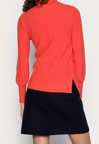 Roter gerippter Pullover mit hohem Kragen und puffierten langen Ärmeln, kombiniert mit einem schwarzen Rock mit seitlichen Schlitzen. Weicher, strukturierten Stoff.