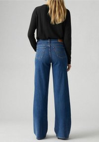 Spodnie dżinsowe damskie z szerokimi nogawkami i wysokim stanem, wykonane ze średnio niebieskiego denimu, z brązową skórzaną łatą z tyłu oraz ozdobione przeszyciami na tylnych kieszeniach.