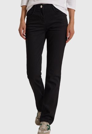 Personne portant un pantalon noir à jambes droites et des baskets beiges avec des accents verts, debout devant un fond uni.
