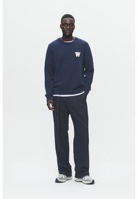 Tumedavarine kudum sweatshirt, millel on kreemivärvi "W" logo rinnal, koos tumedate pükste ja hallide tossudega. Lihtne, juhuslik disain.