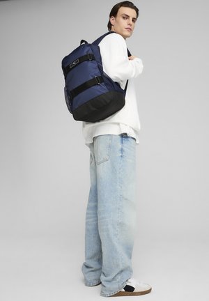Zaino blu navy con base nera, dotato di spallacci regolabili, tasche laterali in rete e due fibbie frontali; materiale tessile con design elegante.