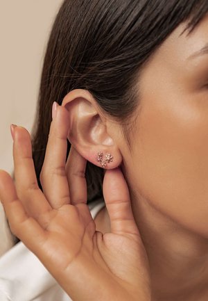 Rose gold flower-shaped stud earrings with intricate detailing, displayed near an ear, accentuated by smooth, straight hair.