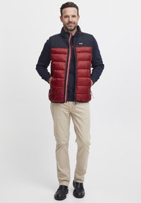 Gilet rembourré rouge avec un col bleu marine, fermeture éclair et poches latérales. Porté sur une chemise à manches longues bleu marine et associé à un pantalon beige.