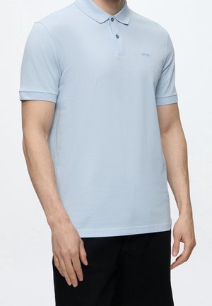 Man die een lichtblauw poloshirt met korte mouwen en twee knopen draagt, en een zwarte broek, staand tegen een effen achtergrond.