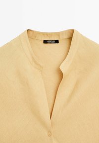 Leinenbluse in hellbeige mit V-Ausschnitt und einer Knopfverschluss. Die Stoffstruktur ist glatt mit einer feinen Webart.