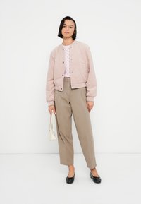 Ροζ ανοιχτό μπουφάν τύπου bomber με κουμπιά τύπου snap, τοπ σε κρεμ χρώμα με σχέδια, μπεζ παντελόνι φαρδιάς γραμμής και μαύρα μπαλαρίνια. Το μοντέλο κρατά μια λευκή τσάντα.