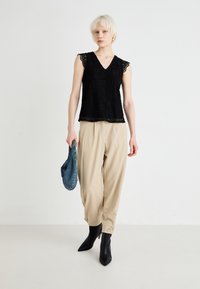 Svart blondetopp med bølgede skuldre, kombinert med løse beige bukser og svarte spissede støvler, holder en blå denimveske.